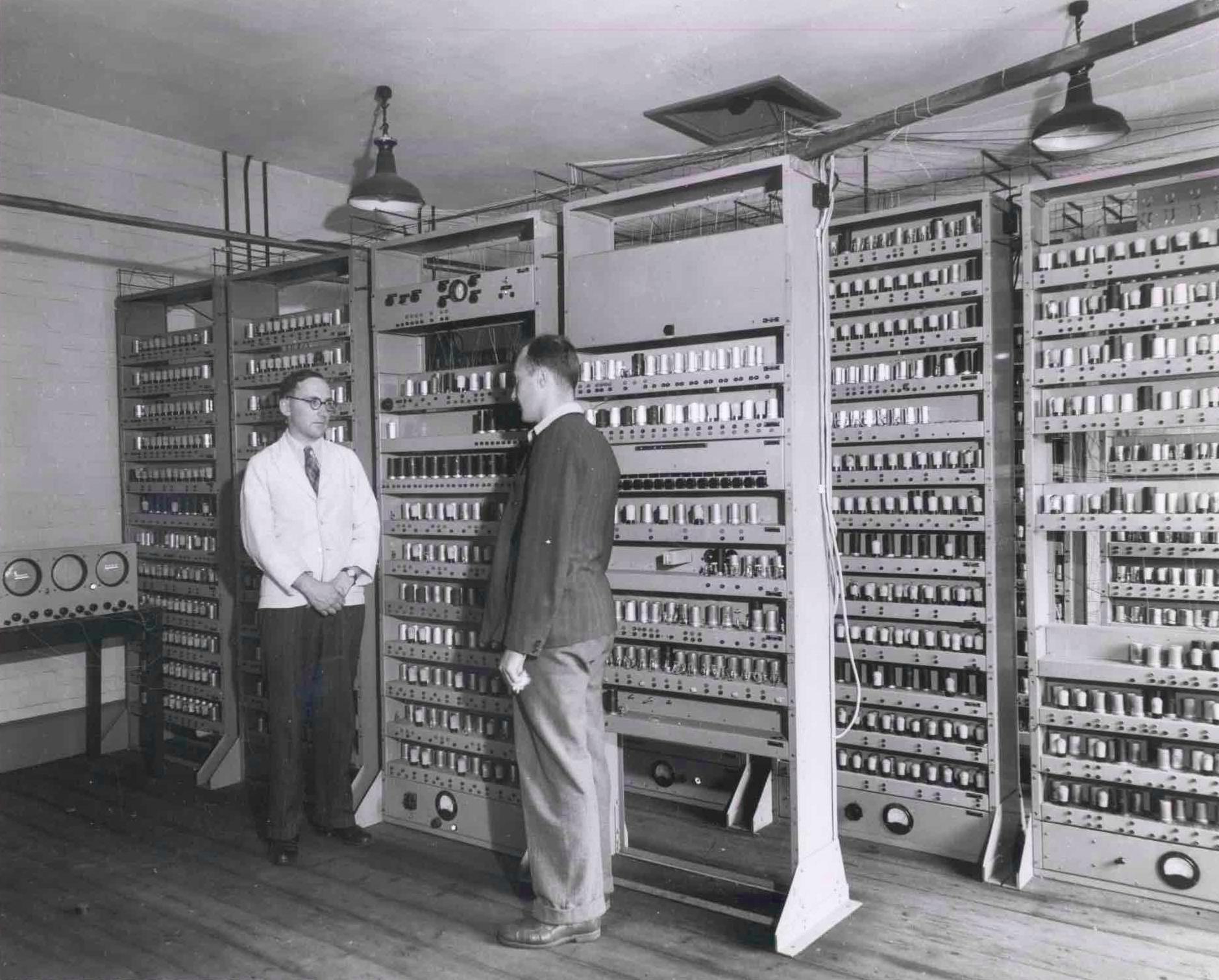 A black-and-white photograph of two men dwarfed by the EDSAC, which fills the room as
            open racks of vacuum tubes standing floor to ceiling in five bays. One man wears a white
            lab coat and faces the camera; the other, in a dark jacket, stands with his back to us
            looking at a rack. Wiring runs overhead across the ceiling, industrial lamps hang above,
            and a bench carrying three circular meters sits at the left edge.